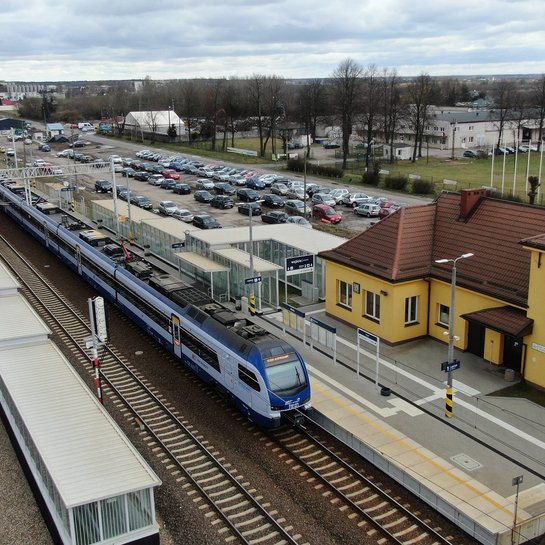 Widok z góry na stację w Warce, widać pociag przy peronie, budynek dworca i parkujące samochody, fot. A.Lewandowski, P.Mieszkowski