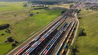 Widok z lotu ptaka na rozległą stację kolejową z wieloma torami i długimi składami pociągów towarowych z kontenerami. Po obu stronach torów znajdują się zielone pola uprawne, a w oddali widać niewielką miejscowość, drogi oraz lasy.