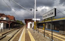 Stacja Brochów. Peron, poczekalnia, tablica z nazwą stacji. Obok przejeżdża pociąg.