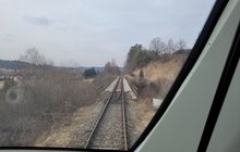 Widok z kabiny pociągu jadącego z Uherzec do przejścia granicznego w Krościenku. Przed pociągiem rozciąga się panorama pobliskich miejscowości – z lewej strony liczne zabudowania, z prawej i w tle las.