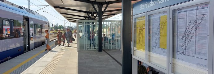 Gablota z informacją o rozkładzie na peronie w Skarżysku-Kamiennej, w tle podróżni i pociąg. fot. Iza Miernikiewicz