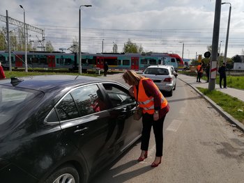 Przedstawiciele PLK SA rozmawiają z kierowcami przed przejazdem kolejowo-drogowym w Łodzi. Na pierwszym planie sznur aut stojących w kierunku przejazdu. Po torach jedzie pociąg. Na chodnikach po obu stronach jezdni stoją piesi; fot. Rafał Wilgusiak