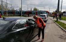Przedstawiciele PLK SA rozmawiają z kierowcami przed przejazdem kolejowo-drogowym w Łodzi. Na pierwszym planie sznur aut stojących w kierunku przejazdu. Po torach jedzie pociąg. Na chodnikach po obu stronach jezdni stoją piesi; fot. Rafał Wilgusiak