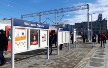Peron z tablicą z nazwą stacji Warszawa Główna, w tle podróżni.