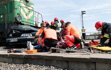 Grupa ratowników udziela pomocy poszkodowanemu kierowcy, w tle lokomotywa i wrak samochodu.