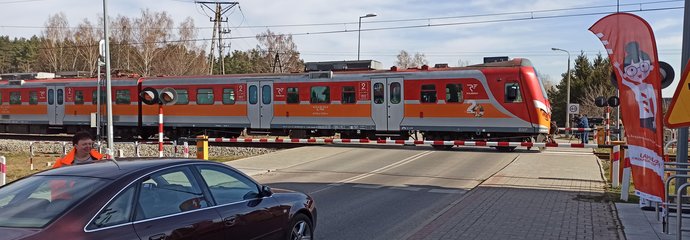 Pracownica PLK rozmawia z kierowcą samochodu przy przejeździe kolejowo-drogowym w ramach akcji edukacyjnej kampanii „Bezpieczny przejazd”. Przed samochodem przejeżdża pociąg. Fot. Katarzyna Głowacka