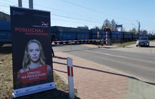 Na pierwszym planie widoczny banner kampanii Bezpieczny przejazd, a w tle pociąg przejeżdżający przez przejazd kolejowy, fot. Katarzyna Głowacka.