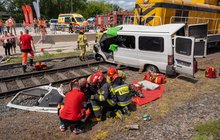 Grupa ratowników udziela pomocy poszkodowanemu kierowcy, w tle lokomotywa i wrak samochodu.