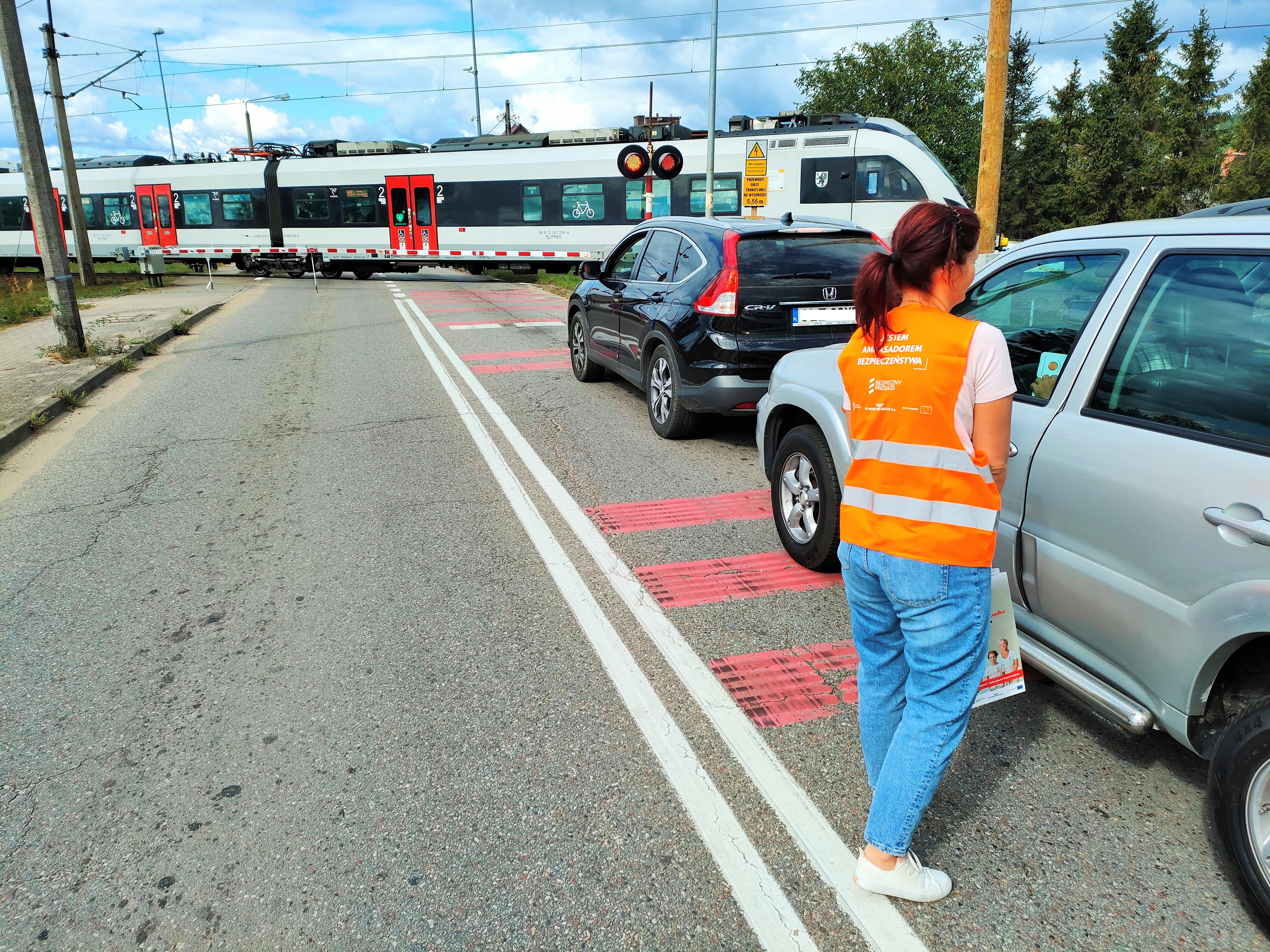 Akcja Bezpieczny Piątek - w miejscowości Mosty. k. Lęborka - samochody przed przejazdem, przedstawiciel PLK rozdaje ulotki. W oddali przejeżdża pociąg fot. Przemysław Zieliński.jpg