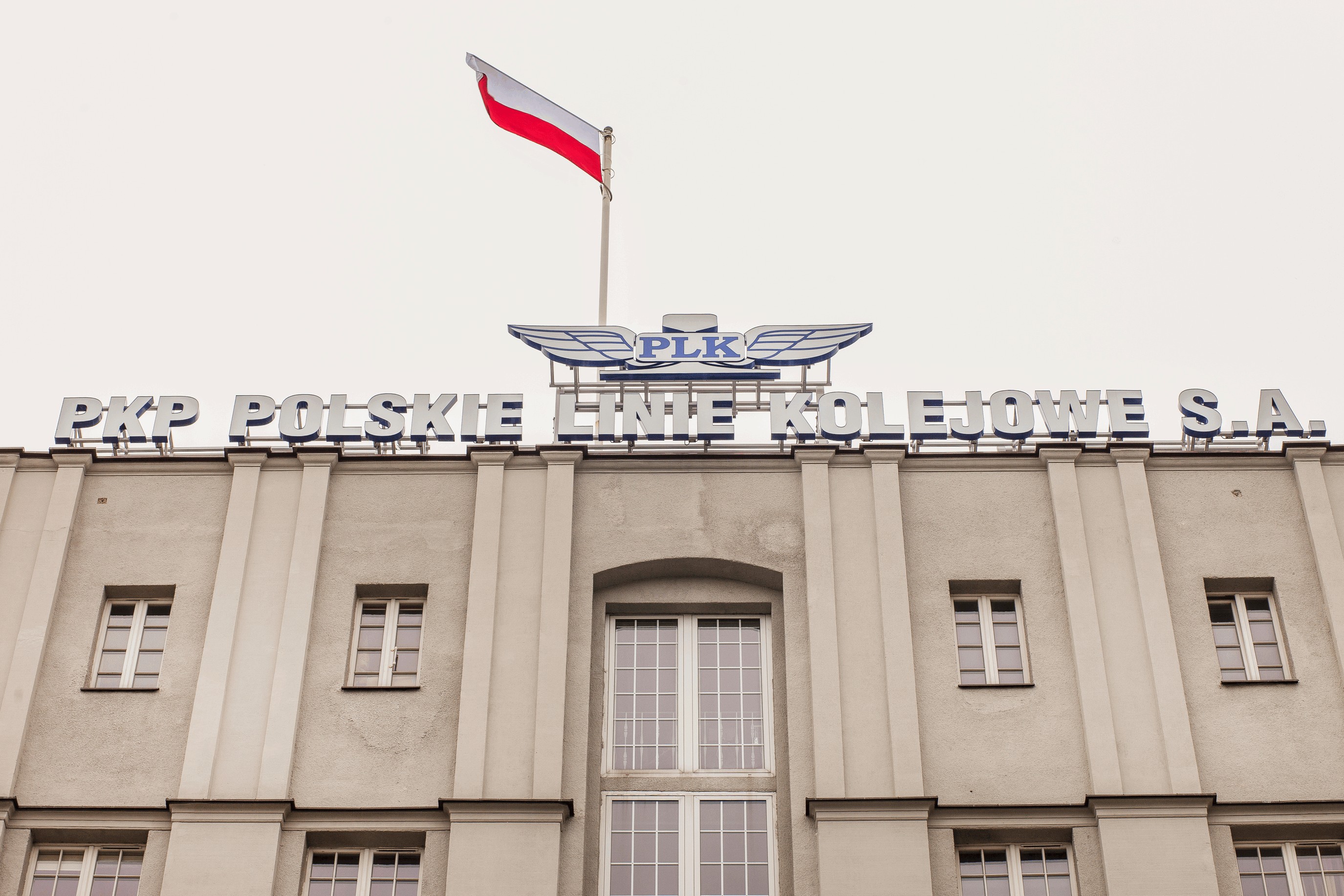 Widok na środkową część budynku gmachu (łącznik), na dachu flaga Polski oraz przestrzenne logo 3D PKP Polskie Linie Kolejowe S.A. na dachu budynku. 